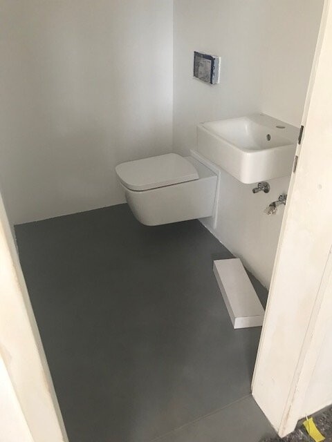 Kleines Bad mit wandhängendem WC, kleinem Wandwaschbecken und grauer Boden.