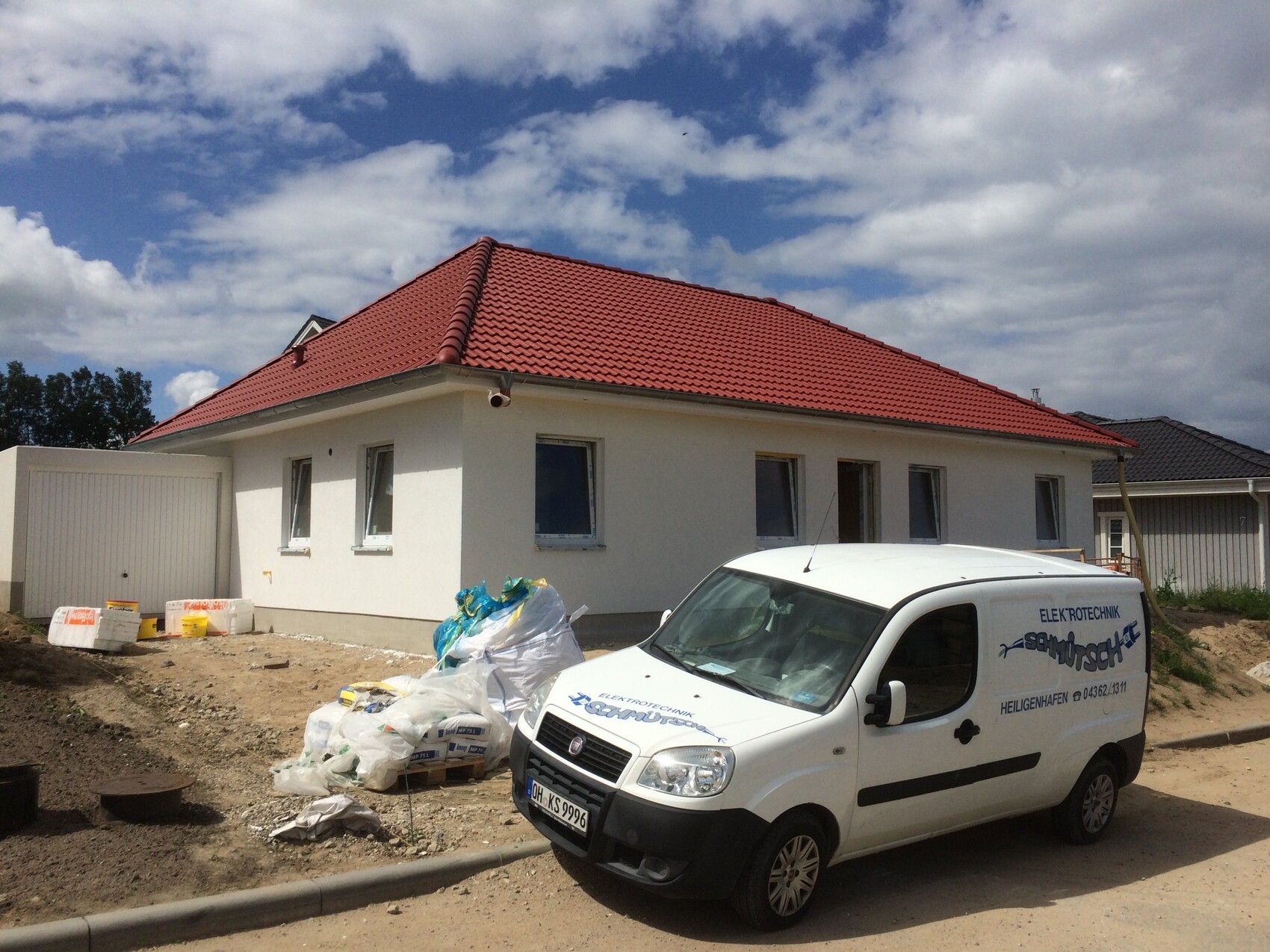 Weißes Einfamilienhaus mit rotem Ziegeldach auf Bauplatz, Baumaterialien und Lieferwagen.