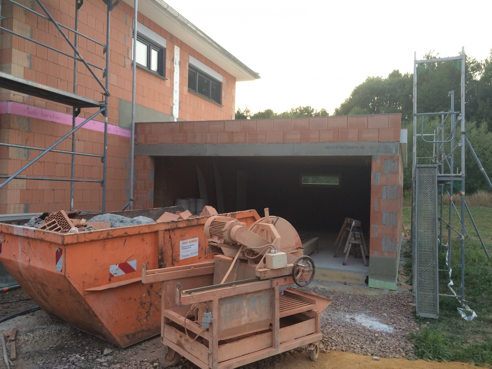 Construction site: red brick house under construction, scaffolding, garage and construction debris container.