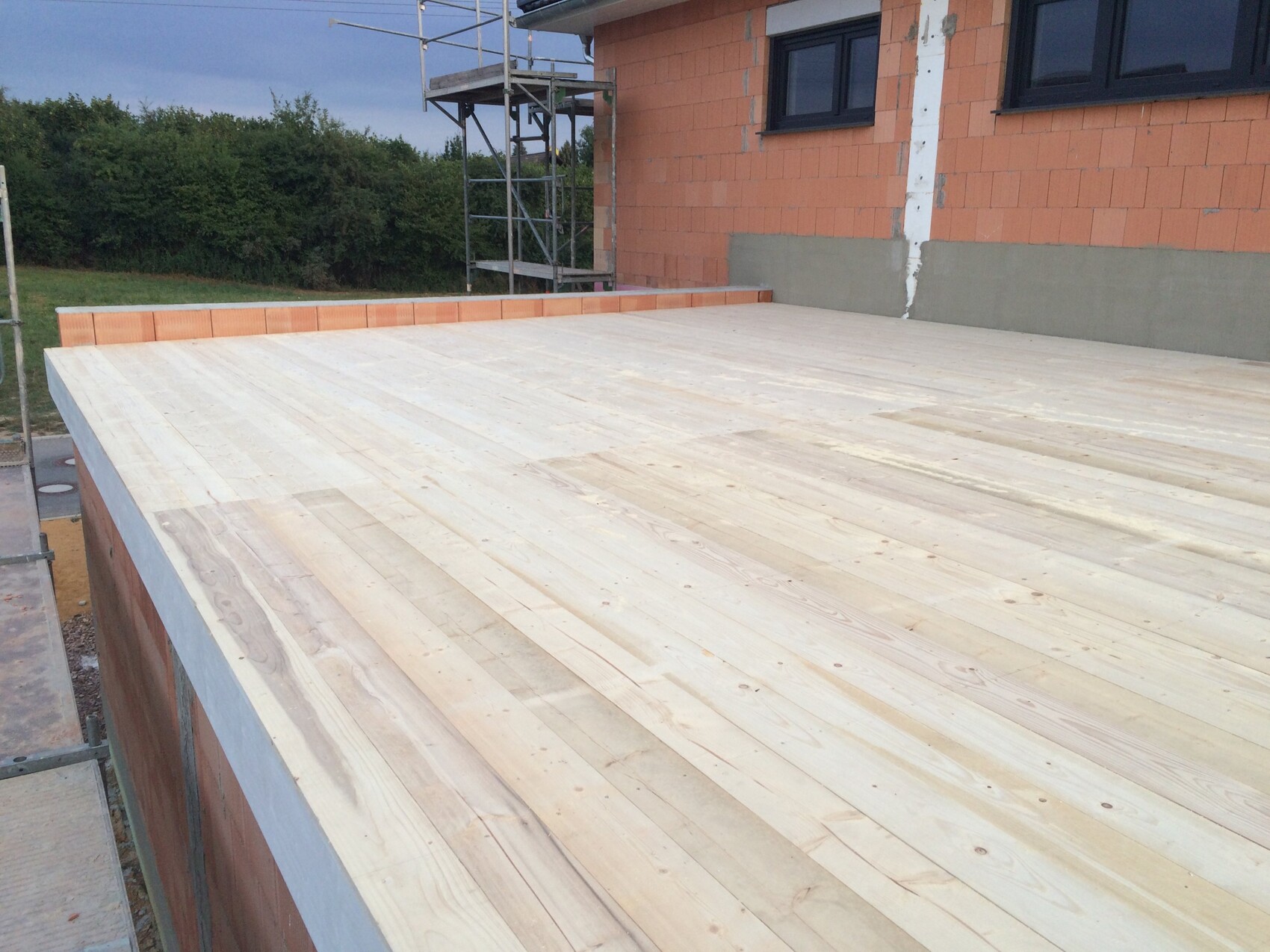 Outdoor area of a construction site with wooden decking, orange brick wall, windows, and scaffolding in the background.