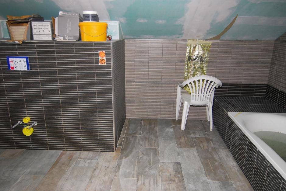 Renovated bathroom with dark tiles, bathtub, white plastic chair, and construction tools.