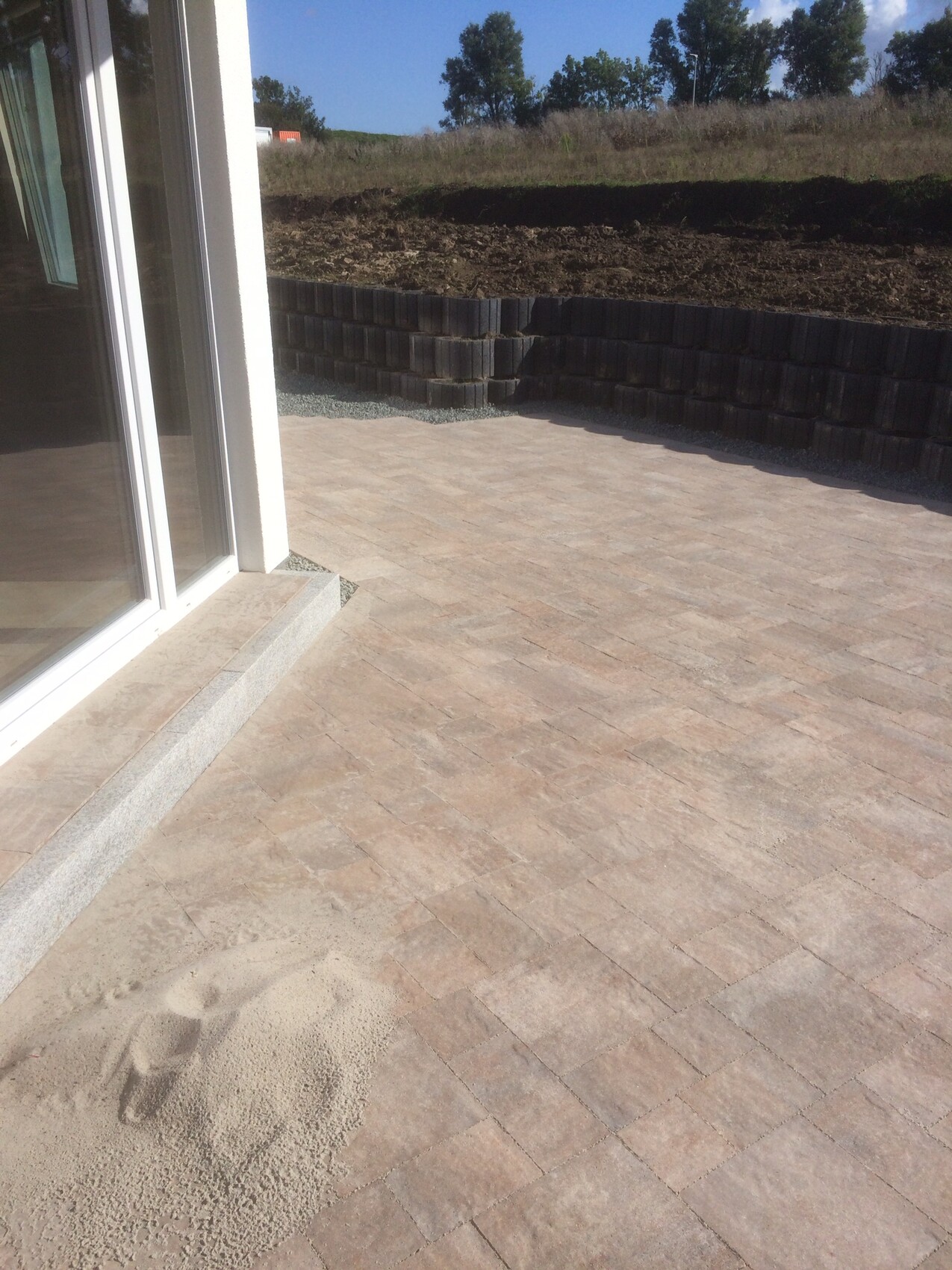 Pflasterte Terrasse mit Glastür; Sandhaufen am Boden, dunkle Betonsteinmauer im Hintergrund.