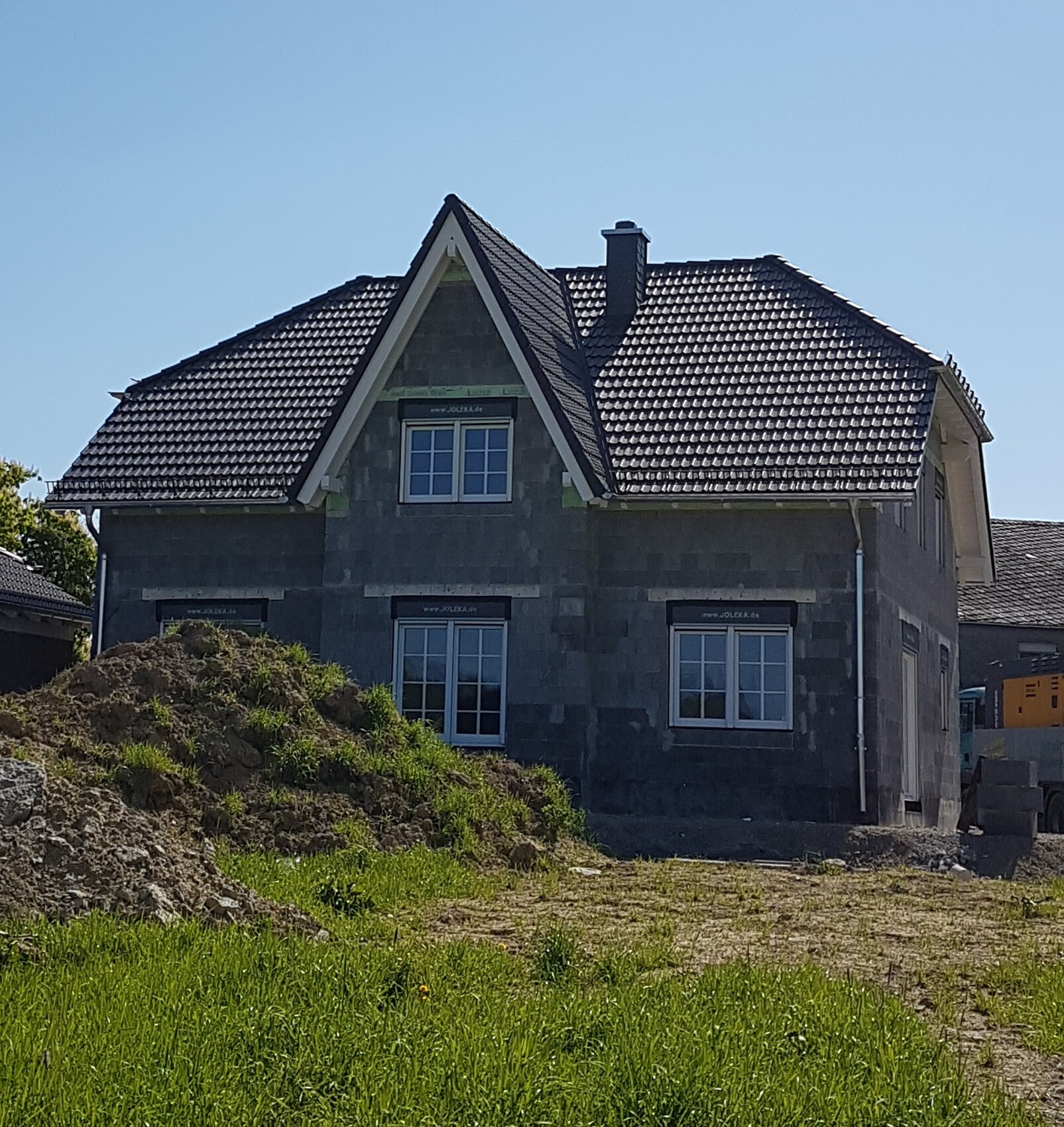House in shell construction stage with dark tiled roof, windows, construction mound in front of green area