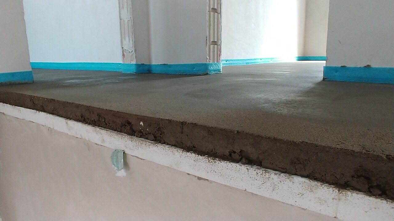 Innenraum mit rohem Betonboden, Türdurchgang sichtbar, blaue Randleisten an Wänden.
