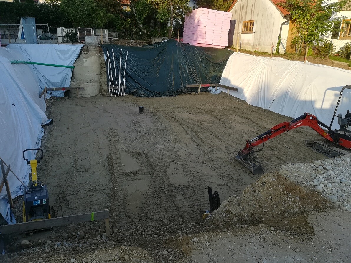 Construction site with earthworks, excavator in foundation trench, tarpaulins and building materials.