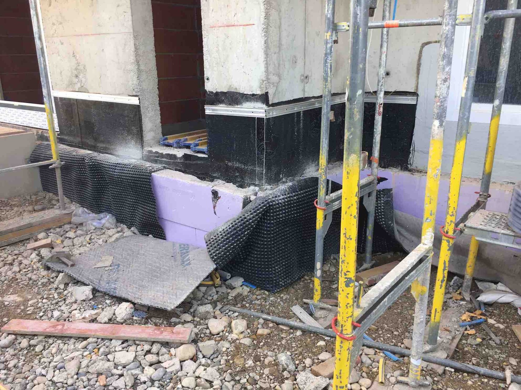 Construction site with foundation, wall construction, scaffolding, insulation boards, and gravel base.