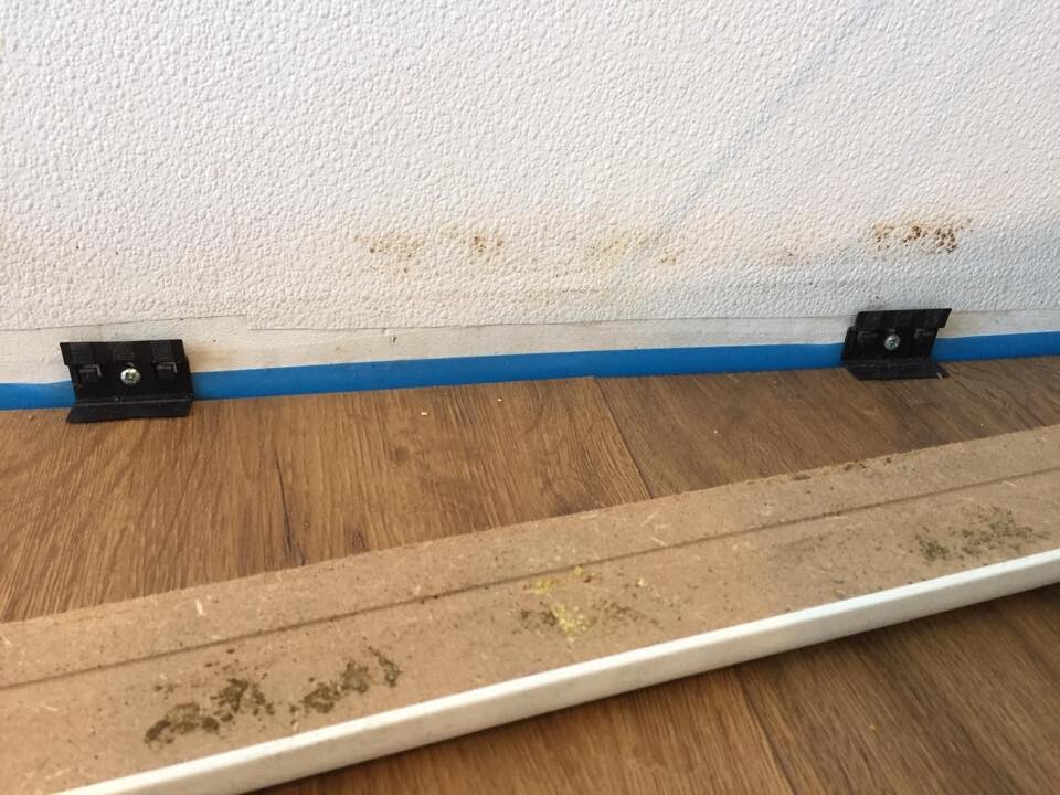 White wall with blue baseboard; two black metal brackets with screws on wooden floor.