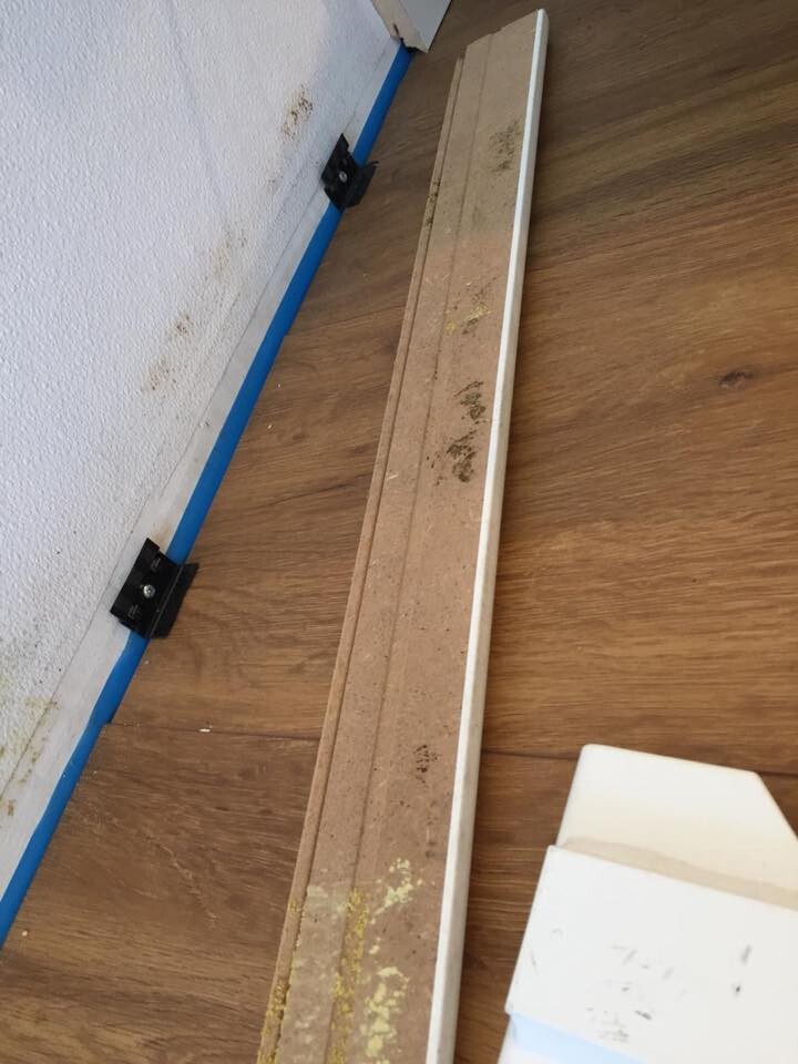 Long wooden baseboard on laminate floor next to white wall; blue painter’s tape and black brackets.
