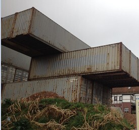 Rostige Containerstapel auf Bauplatz mit Gras und Haus im Hintergrund.