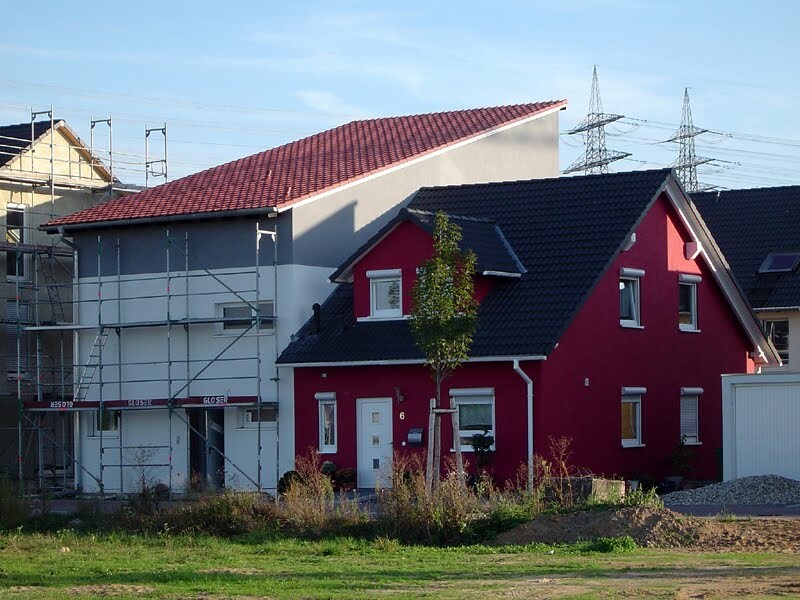 Red detached house in the foreground, building site with scaffolding on the left, green meadow in front.