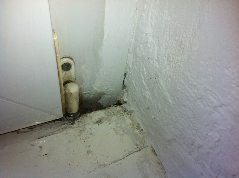 Corner of a room with white walls, damp floor, and a pipe-like drain.