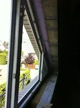 Offener Fensterrahmen im unfertigen Innenraum mit Blick nach draußen auf Garten und Häuser.