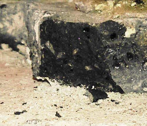 Burnt black block made of stone/brick in front of a rough stone wall, sooty and dusty.