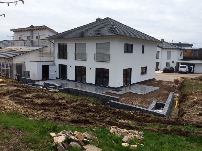 Modern white single-family house with gray roof, terrace; construction work and soil heaps in the garden.