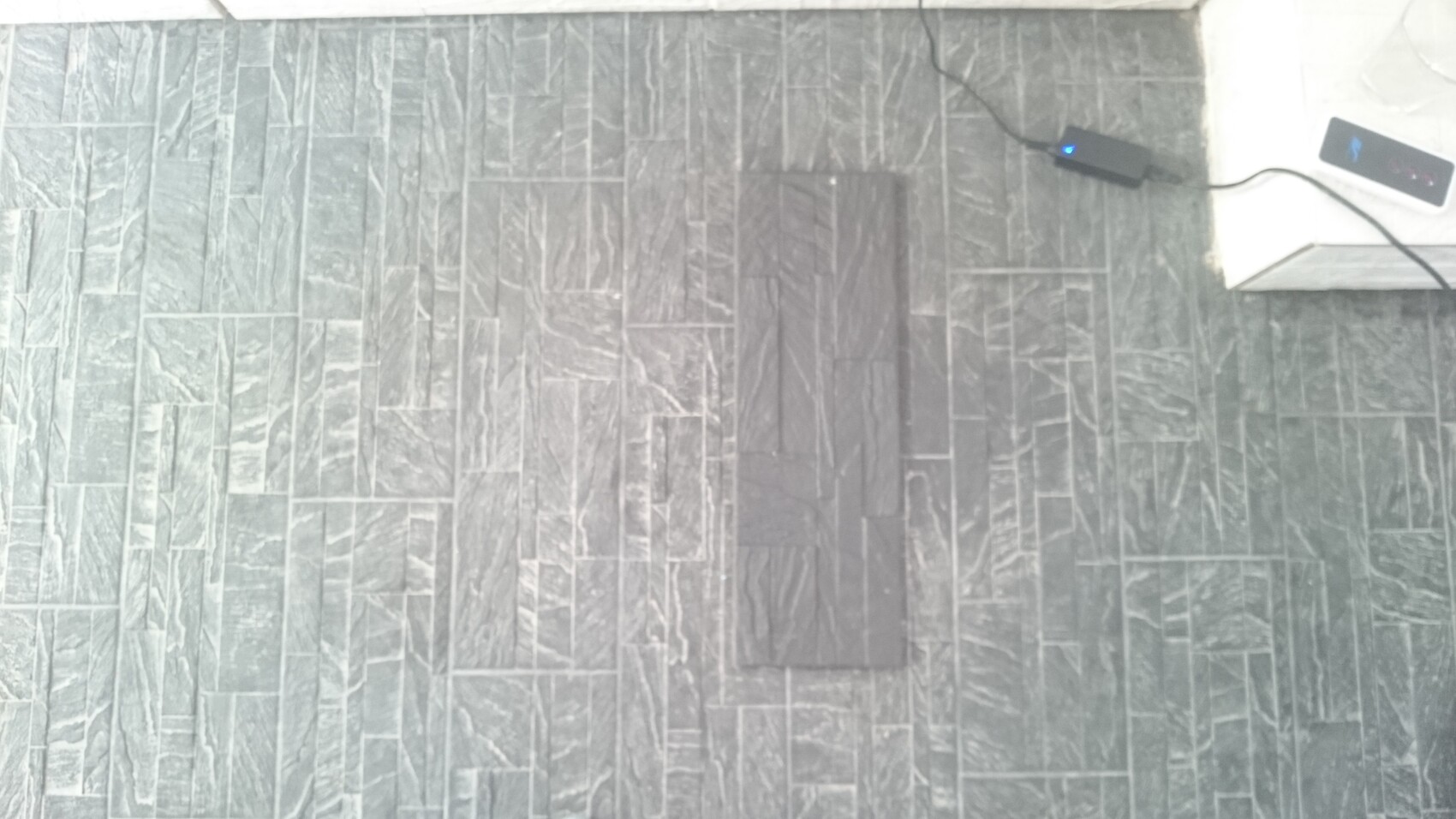Gray patterned tiled floor; in the center a dark rectangular fabric, right side power adapter.