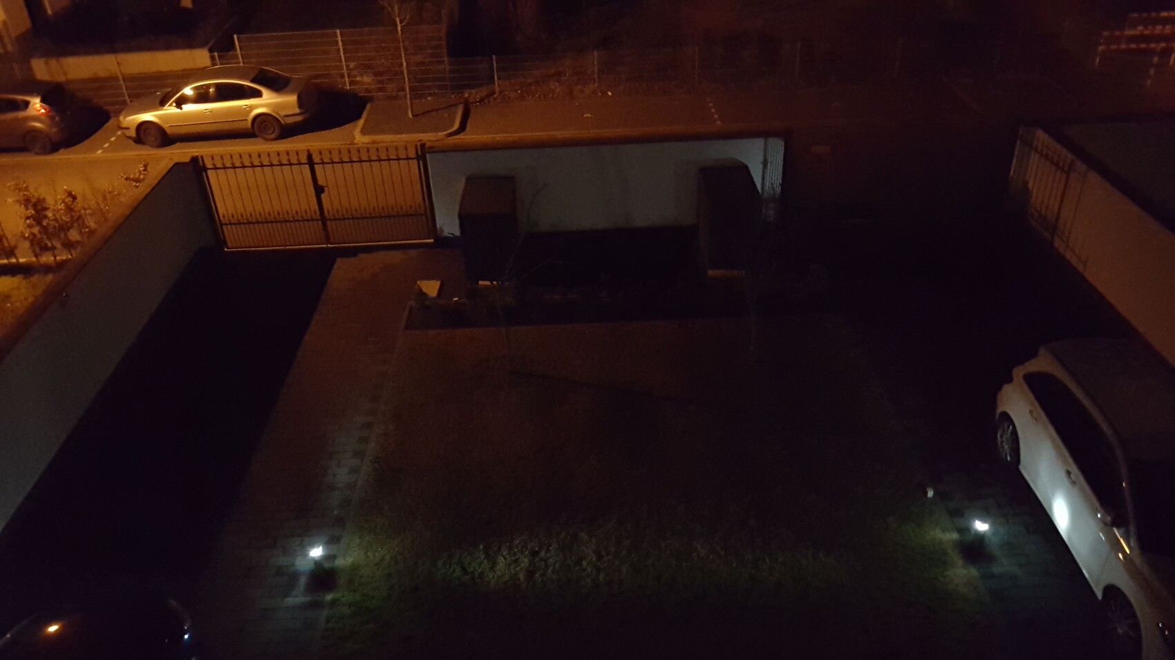 Night view of a courtyard with gate, fence, and parked cars.