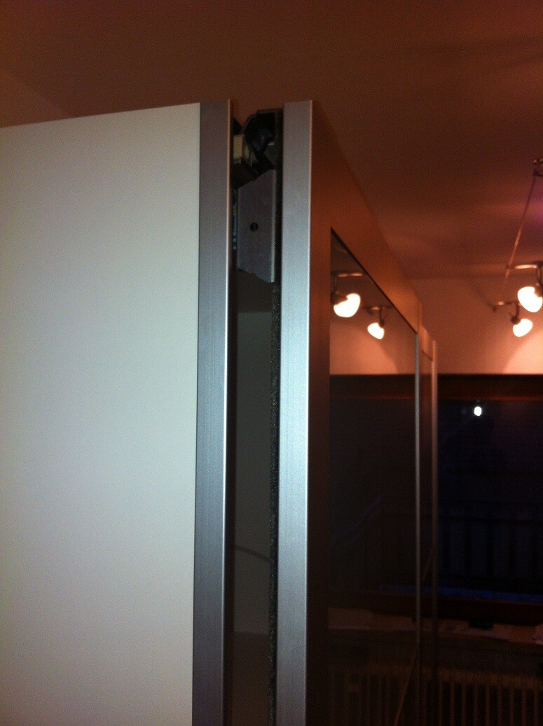 Open door of a cabinet with silver frame, glass panels and ceiling lights in the background