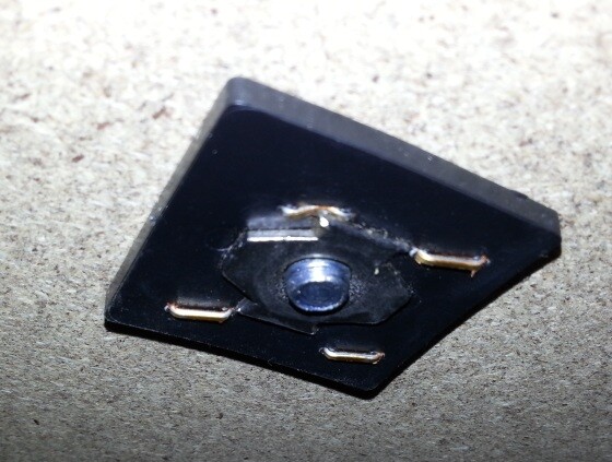 Small black mounting plate with central opening and four metal pins on rough concrete floor.