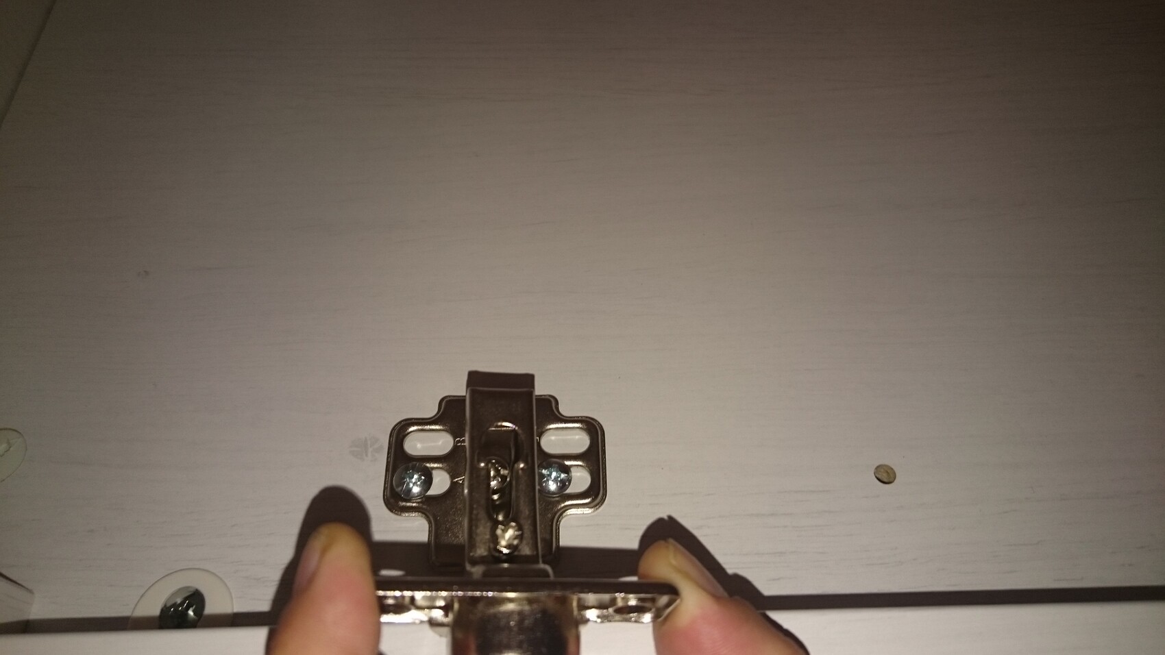 Close-up of a door hinge with screws, held by a hand on a wooden door.