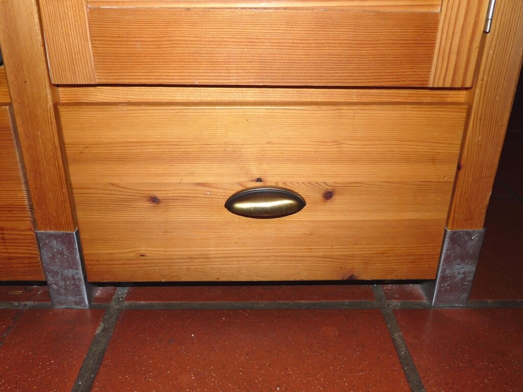 Wood drawer of a lower kitchen cabinet with metal foot caps in front of tiled floor.
