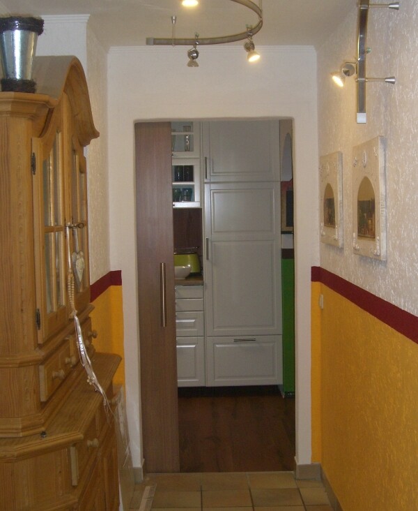 Narrow corridor with wooden cabinet on the left, view through door into kitchen with gray kitchen cabinets.