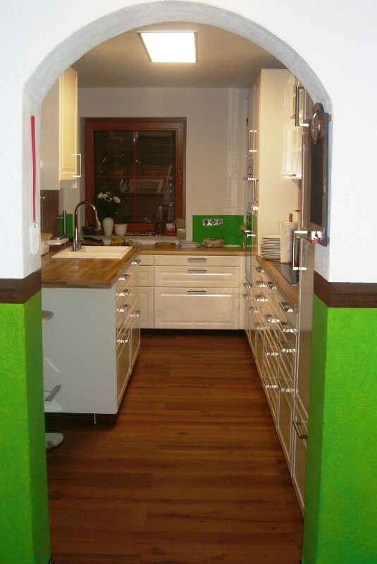Kitchen through arched opening: white cabinets, wooden floor, green accents, sink.