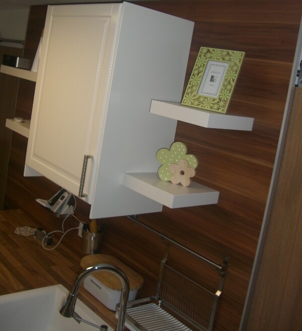 White kitchen cabinets on wooden wall, floating shelves with floral decoration and frames; sink visible.
