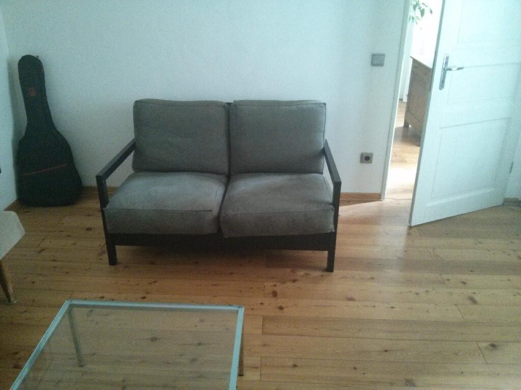 Living room with gray sofa on wooden floor, glass table in the foreground and guitar case on the wall.