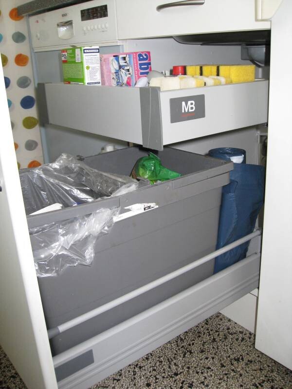 Open kitchen cabinet with pull-out drawers, cleaning supplies, garbage bags, and cleaning utensils.