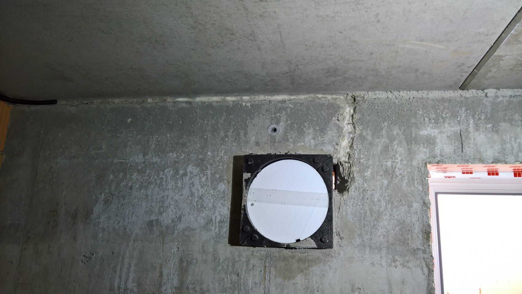 Basement wall with round ventilation opening in a black square frame; window on the right side of the image