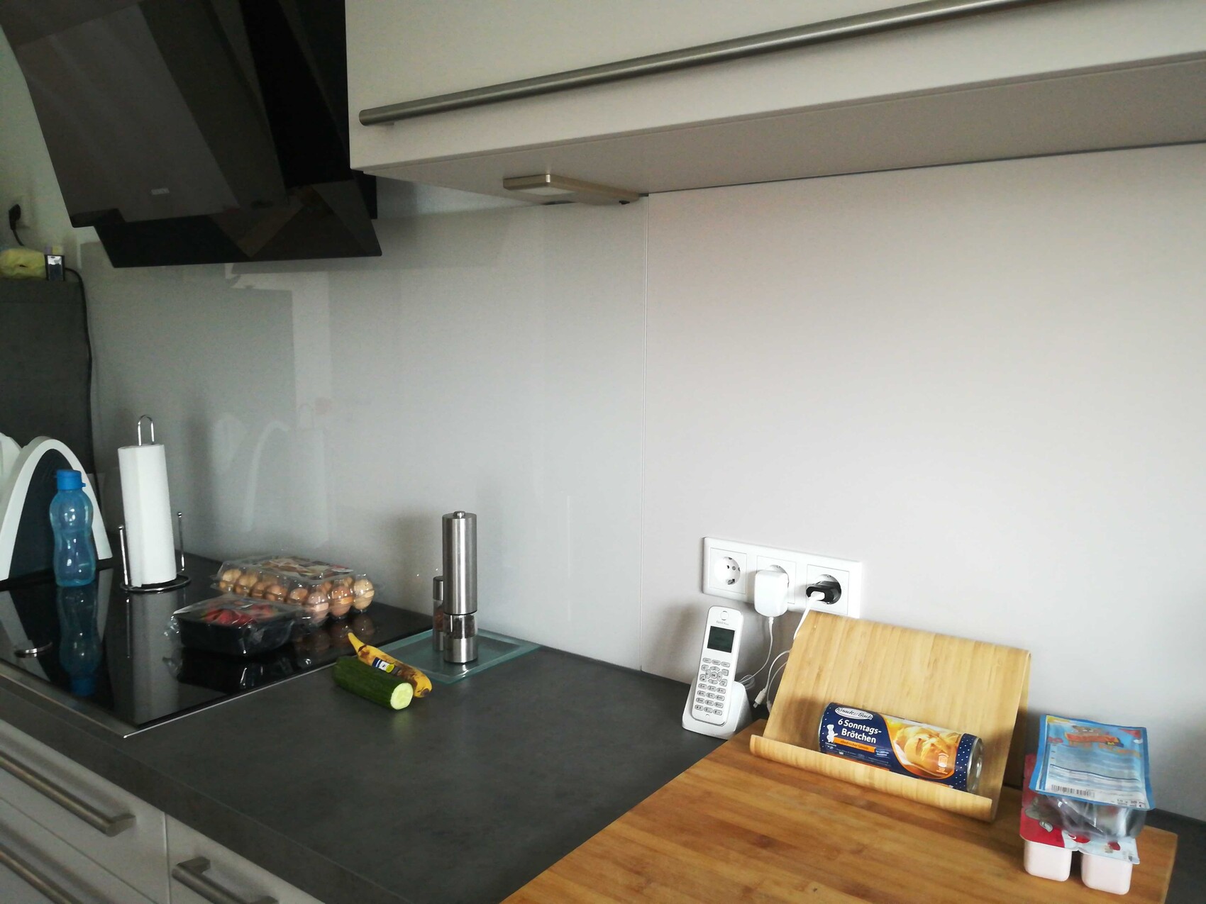 Modern kitchen unit with glass backsplash, countertop, egg carton, cucumber, kitchen roll, and phone.