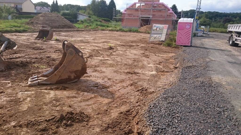 Construction site with excavator bucket, building plot, and portable toilet in the background.
