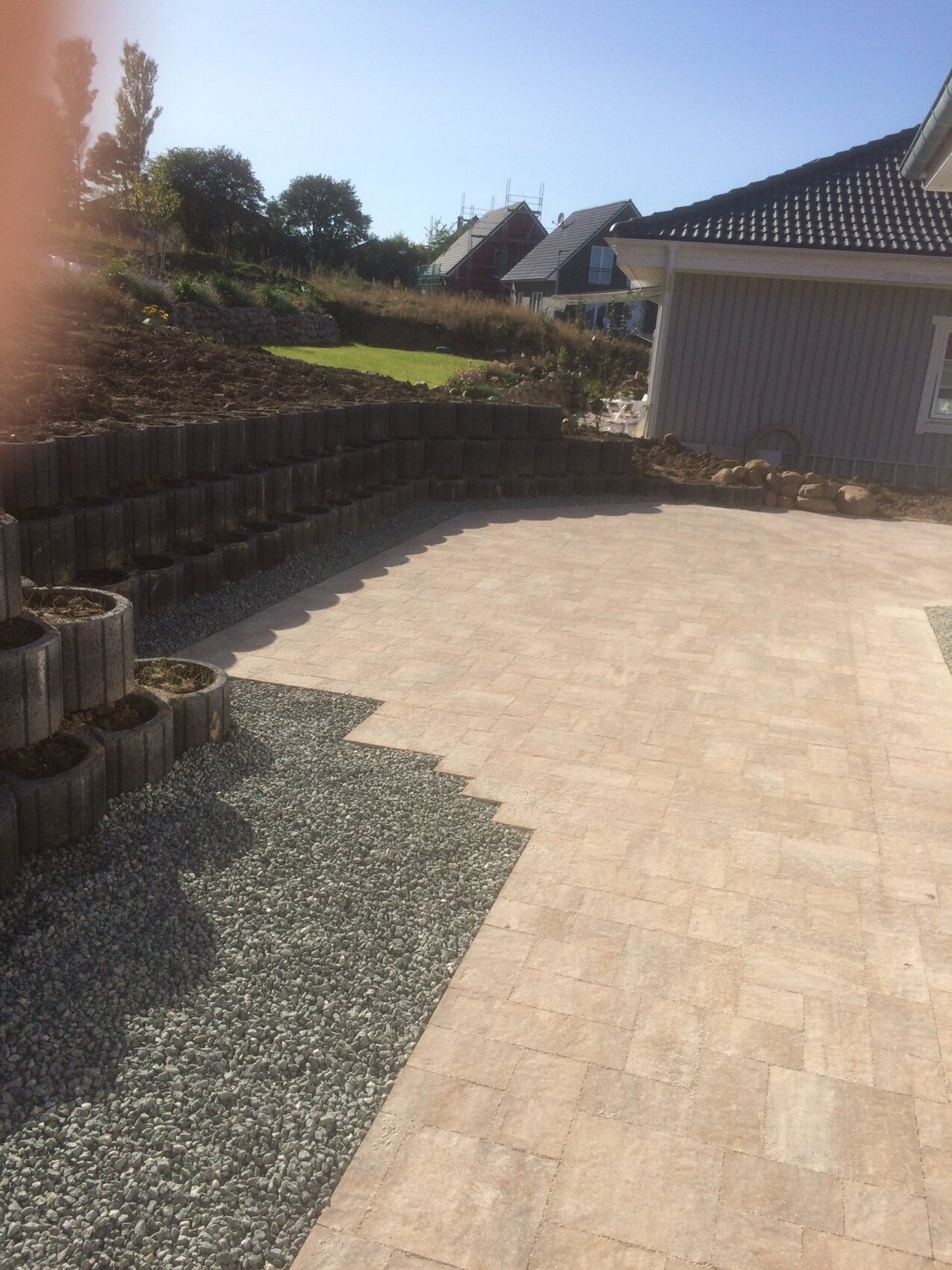 New terrace area with beige paving stones, gravel path and retaining wall made of concrete blocks in front of house.