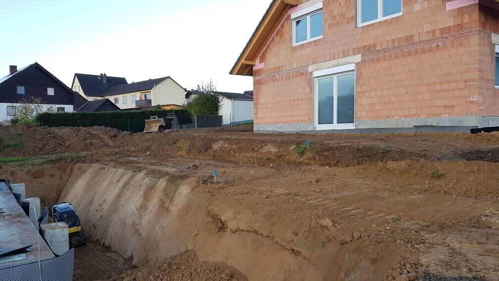 Construction site: deep excavation next to an unfinished single-family house; excavator on the left.