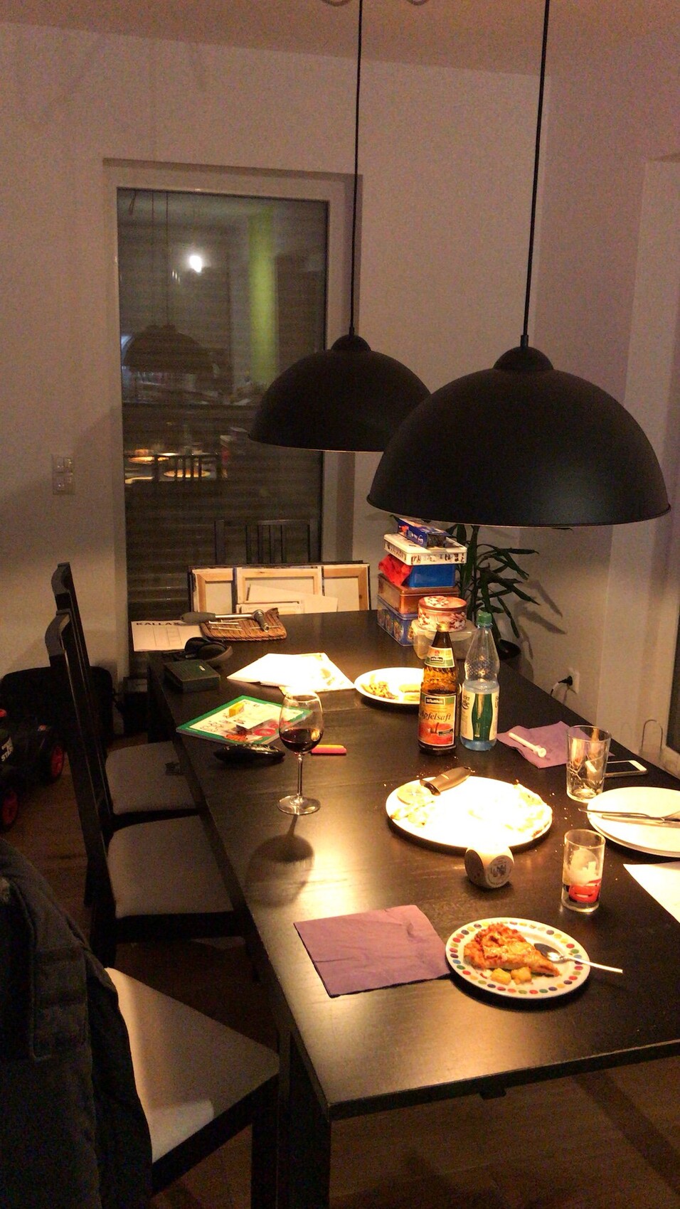 Dining table with plates, red wine in glass, beer bottle, and water; two black hanging lamps.