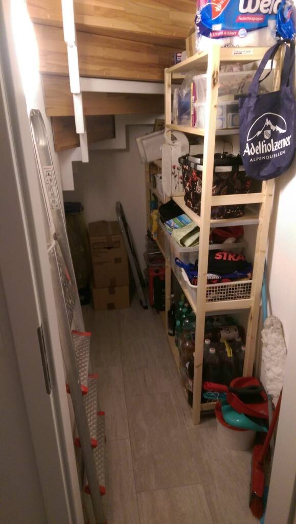 Small storage room with wooden shelves full of bottles, crates, cleaning supplies, ladder.