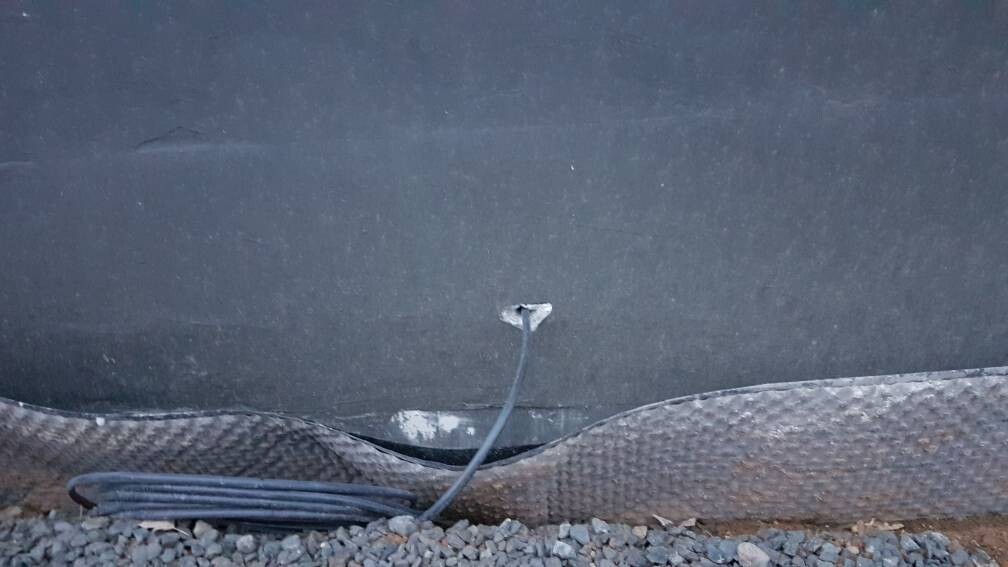 Large protective membrane lying on gravel in front of a wall; next to it a hose reel and cables.