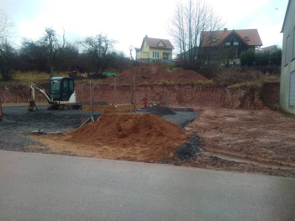 Small construction site with mini excavator, piles of sand and gravel, marked-off building area, houses in the background.