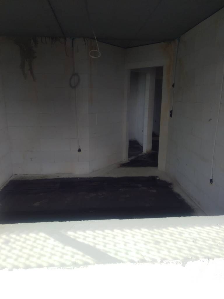 Rohbau eines Raums mit freiliegenden Kabeln, Ringen von der Decke, Türdurchgang und dunklem Boden.