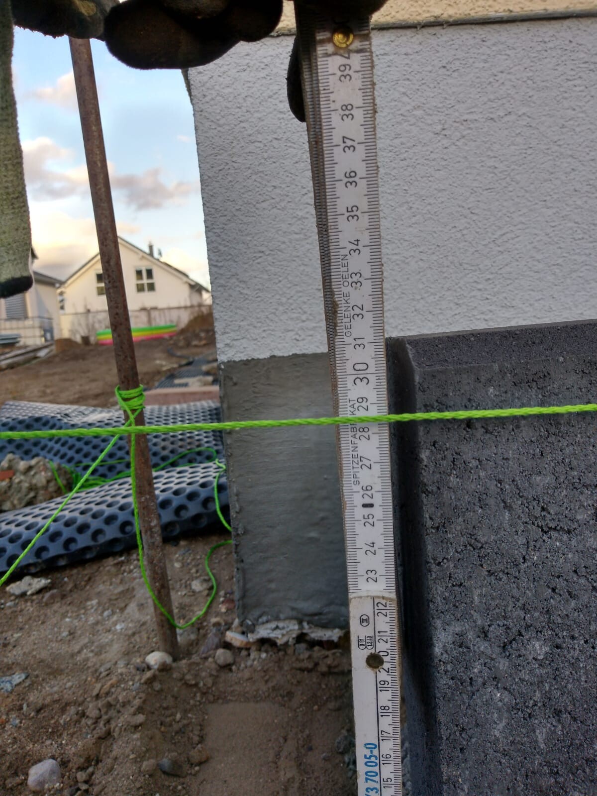 Construction site: measuring rod against concrete wall, green string stretched, ground and building visible.