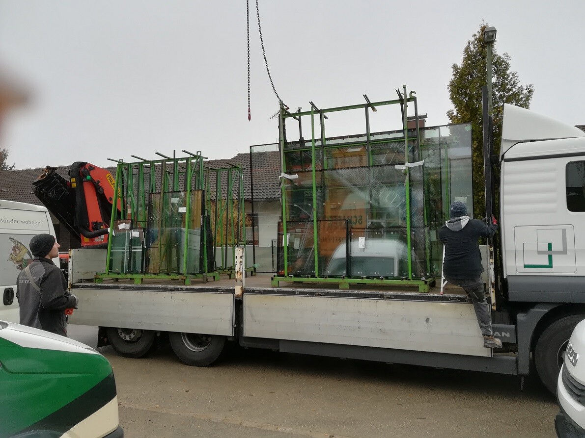 Truck with large glass panels in green frames, crane lifting glass panels, two men watching.