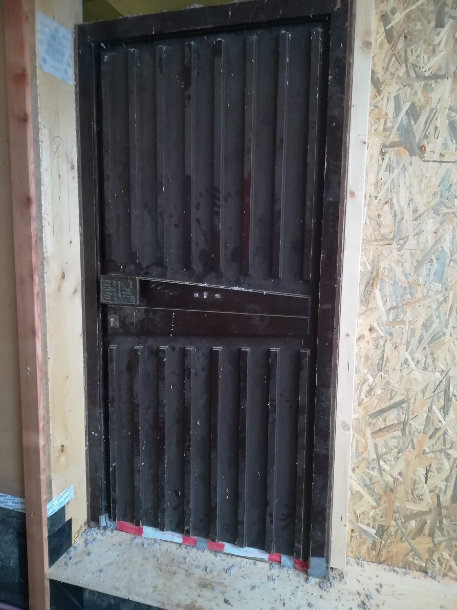 Dark door with vertical wooden slats in construction environment; OSB cladding visible.