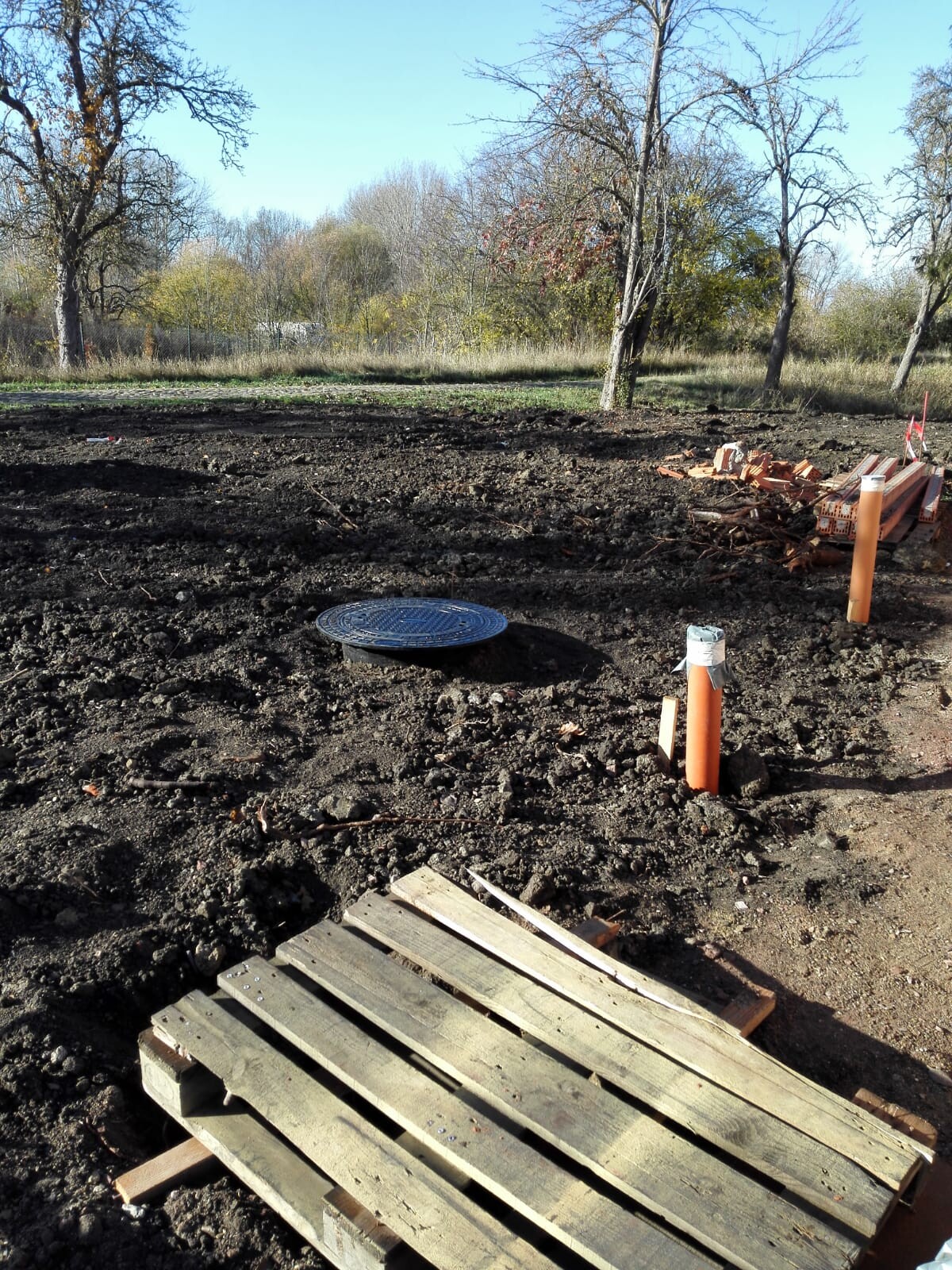 Construction site outdoors: earthworks, wooden pallets, orange pipes, rubble, trees in the background.