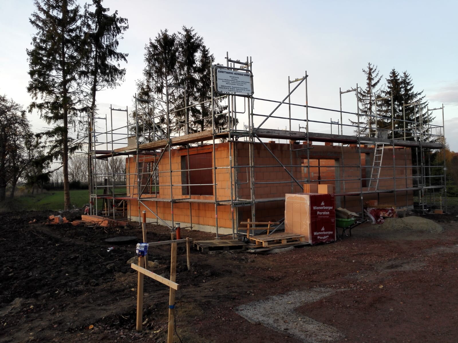 Small house made of orange bricks under construction, surrounded by scaffolding, building materials next to it, trees in the background.