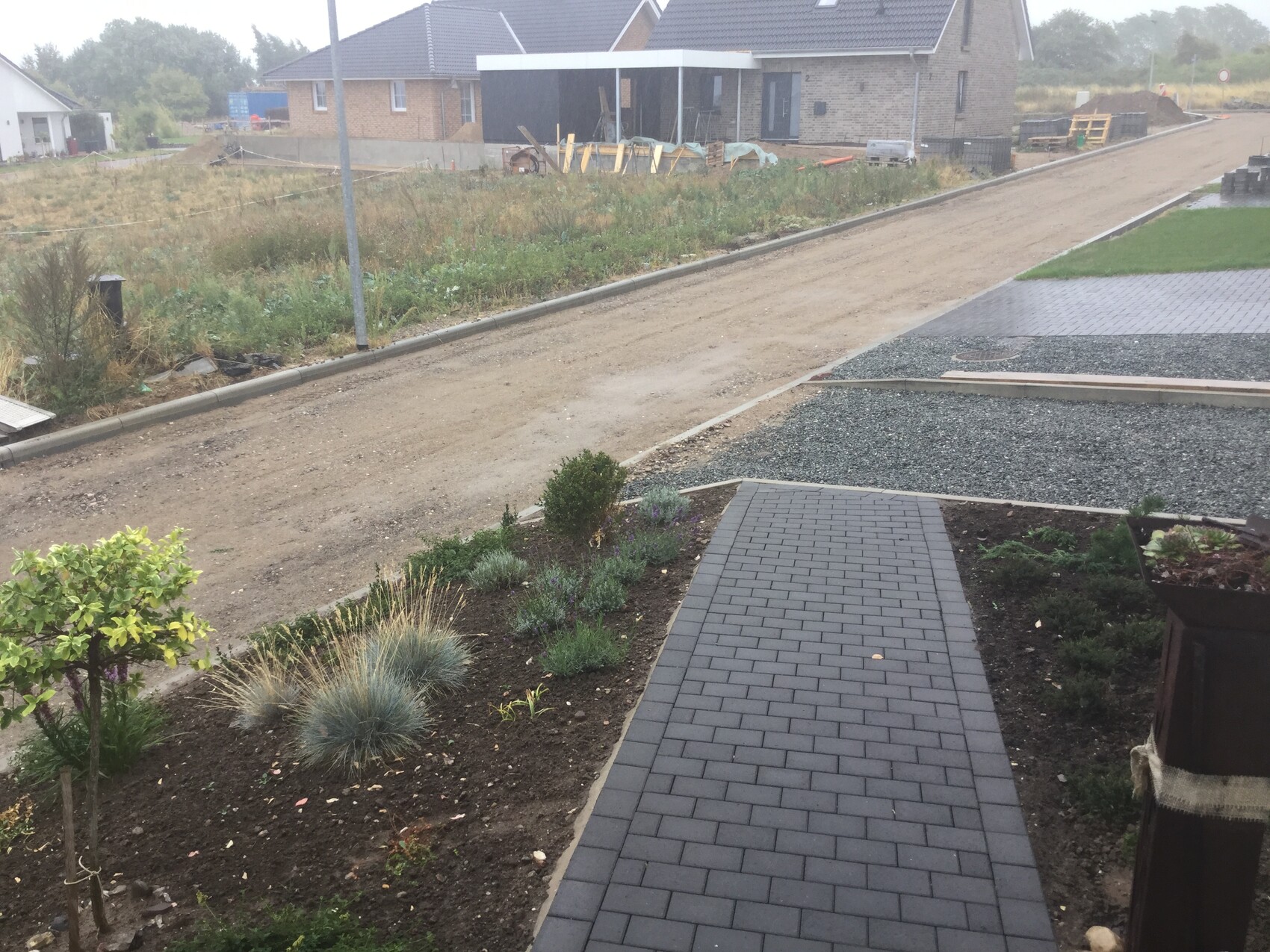 Unpaved driveway in front of new house; garden path, flower beds, and shrubs.