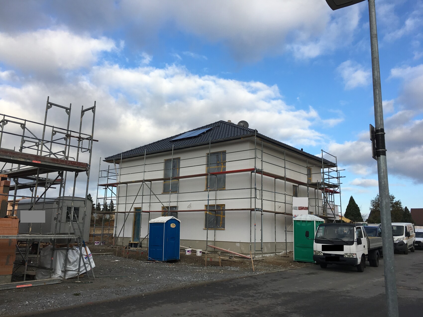 Two-story house under construction with scaffolding; construction container, blue portable toilet, and parked vehicles.