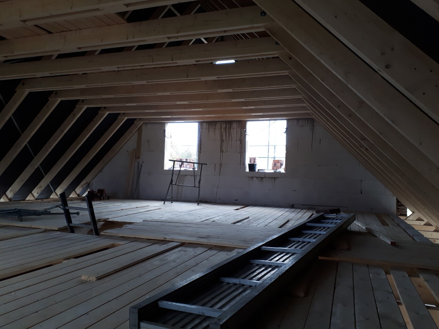 Unfinished attic with visible wooden framework, building materials, ladder, and two windows.