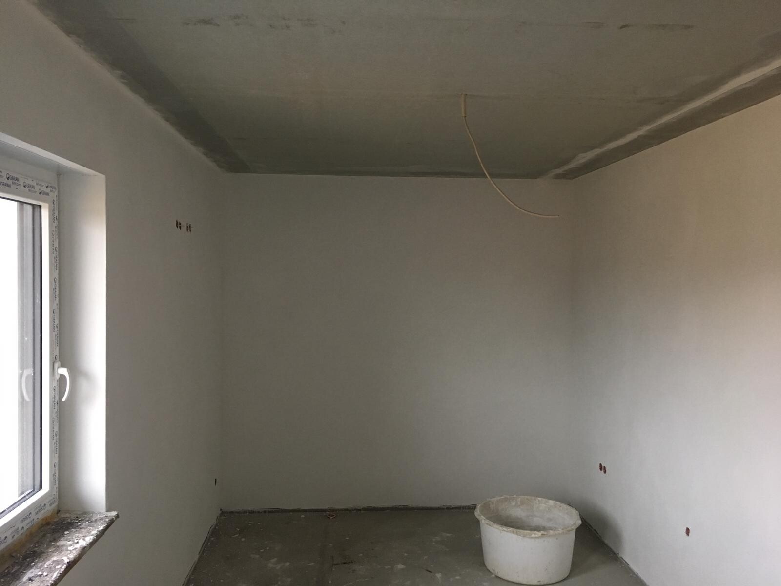 Unfinished renovation room with window on the left, loose cables hanging from the ceiling and a bucket on the floor.