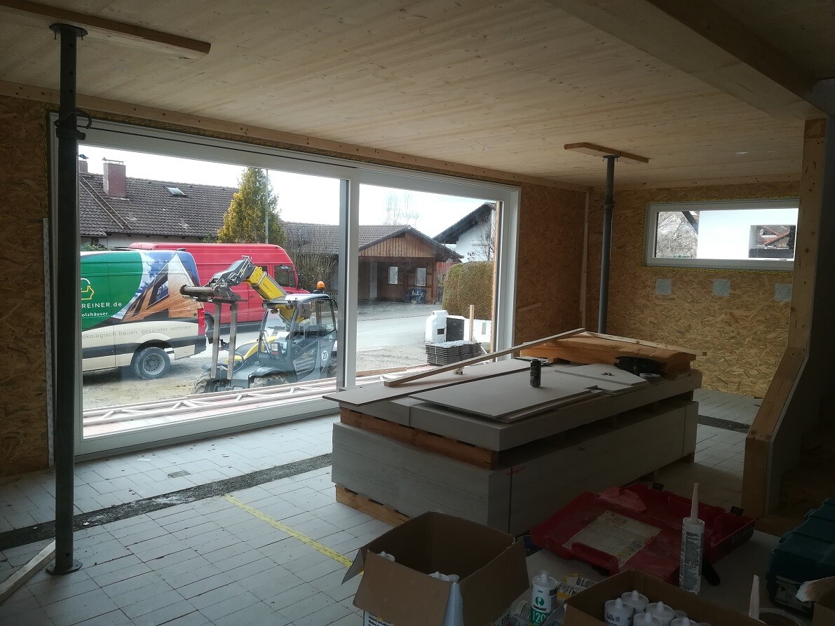 Innenraum einer Baustelle: Stapel Bauplatten, Werkzeuge, Fensterfront mit Baufahrzeugen draußen.