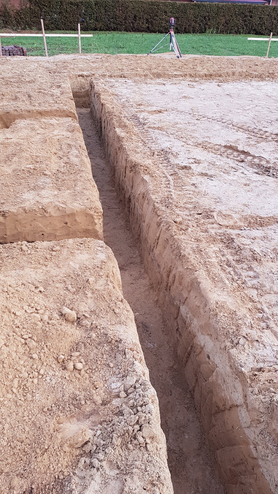 Baugrube mit seitlichen Wänden, Vermessungsstativ im Hintergrund auf grünem Gelände.